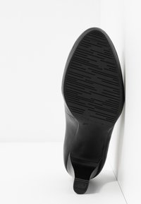 Chaussure noire à talons hauts positionnée semelle vers le haut, montrant une semelle extérieure en caoutchouc texturée avec des rainures décoratives et un petit talon.