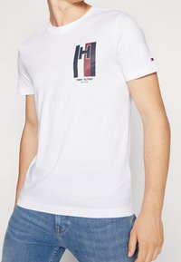 Bílý bavlněný tričko s kulatým výstřihem, se barevným logem na hrudi a krátkými rukávy. Standardní střih, hladká textura.