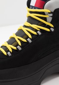 Černé semišové tenisky se žlutými tkaničkami a šedým kováním. Mají strukturovanou černou podrážku a nášivku s logem v červené, bílé a modré barvě.