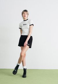 White short-sleeve t-shirt with black ringer details and "Dickies" logo, paired with black shorts and black combat boots, on green floor.