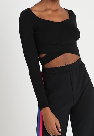 Kvinde iført en sort langærmet crop top med omslagsdetalje og sorte træningsbukser med blå og lyserøde striber i siden.