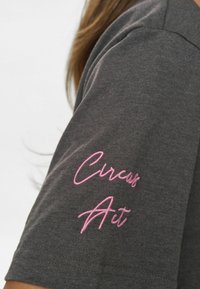 Siva kratka majica s kratkimi rokavi z mehko teksturo; na levem rokavu ima rožnato vezeno besedilo "Circus Art".