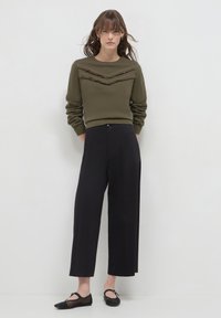 Giovane donna che indossa un maglione oliva con strisce a zigzag frangiate e pantaloni neri a gamba larga cropped, in piedi davanti a uno sfondo bianco.