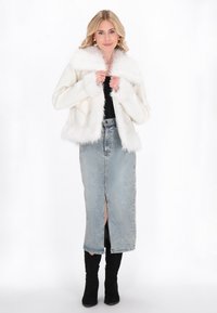 Blonde vrouw in een witte jas met nepbont, een zwart topje, een blauwe denim midi rok met een split aan de voorkant en zwarte knielaarsen.