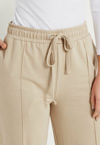 Pantalones de chándal beige con cintura elástica y cordón ajustable. Cuenta con bolsillos laterales y detalles de costura vertical. Textura suave.