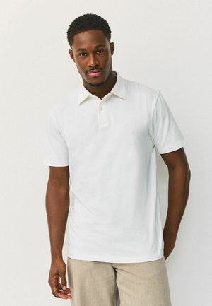 Man wearing a plain white short-sleeve polo shirt and light beige pants, standing against a white background.