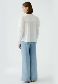 White long-sleeve blouse with puffed shoulders, gathered back, and button closure. Paired with light blue wide-leg jeans.