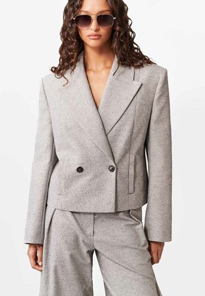 Femeie purtând un sacou gri deschis texturat, cu două rânduri de nasturi, și pantaloni asortați, croiți, cu părul lung și creț și ochelari de soare închiși la culoare.