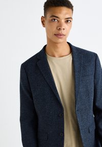 Blazer navy realizzato in tessuto testurizzato con due bottoni, revers a scansione e taschino sul petto, abbinato a una t-shirt beige.