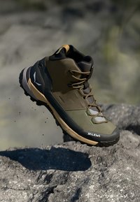 Buty trekkingowe z brązowo-zielonym syntetycznym wierzchem, czarnymi akcentami, teksturowaną gumową podeszwą, szarymi sznurówkami i logo po boku.