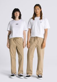 Hvide bomulds T-shirts med Vans-logo, parret med beige drawstring-bukser og sorte Vans-sko på to modeller, der står side om side.