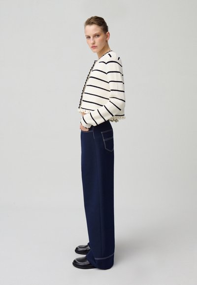 Cardigan en tricot rayé crème et marine, design court avec ourlet à franges. Pantalon large marine avec poches latérales et chaussures noires avec bord blanc.