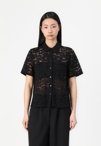 Chemise noire en dentelle à manches courtes avec un col, ornée de motifs floraux, fermeture à boutons à l'avant et texture semi-transparente.