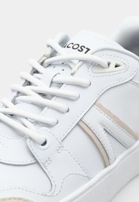 Fehér bőrből készült sportcipő sima felülettel, bézs kiegészítőkkel, fehér fűzőkkel és texturált talppal. Lacoste logó a nyelvén.