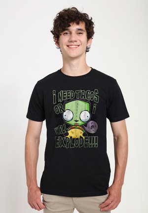 Giovane sorridente con capelli ricci che indossa una maglietta nera con un personaggio dei cartoni animati verde che tiene un taco, con testo relativo alla necessità di tacos.