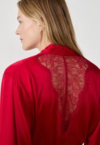 Camicia di seta rossa con pannello posteriore in pizzo caratterizzato da motivi floreali, bordi ondulati e una texture liscia. Maniche lunghe e design con colletto classico.