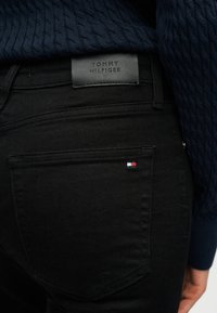 Κοντινό πλάνο μαύρων τζιν Tommy Hilfiger που δείχνουν την πίσω τσέπη με μικρή σημαία του brand και δερμάτινο σήμα στο ζωνάρι.