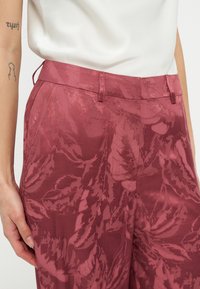 Κοντινό πλάνο ενός ατόμου που φοράει ψηλόμεσα μαρόν παντελόνια με ανάγλυφα σχέδια φύλλων και λευκό τοπ που είναι μαζεμένο μέσα.