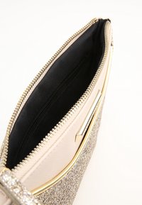 Beige kuvertväska med glittriga silverdetaljer, gulddragkedja och en svart inredning. Har en elegant, rektangulär form och texturerade ytor.