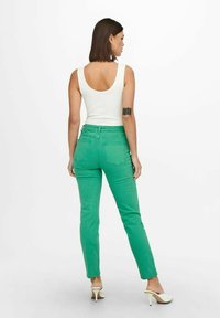 Green denim jeans with a slim fit, featuring back pockets and a mid-rise waist, complemented by a white tank top and high-heeled shoes.