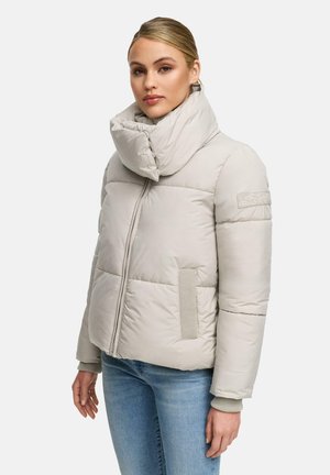 Woman wearing a light beige padded jacket with a high, padded collar and blue jeans, standing against a plain white background.