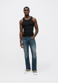 Jovem em pé com uma mão no bolso, vestindo uma camiseta preta justa sem mangas, jeans azuis e sapatilhas cinzas, contra um fundo simples.