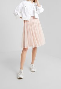 Mujer vistiendo una chaqueta corta blanca, una blusa blanca texturizada, una falda de tul rosa de longitud a la rodilla y zapatillas blancas, de pie sobre un fondo claro y liso.