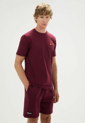 Junger Mann mit kurzen blonden Haaren, der ein passendes burgunderrotes T-Shirt und Shorts mit kleinen Logos trägt, steht mit einer Hand in der Tasche.