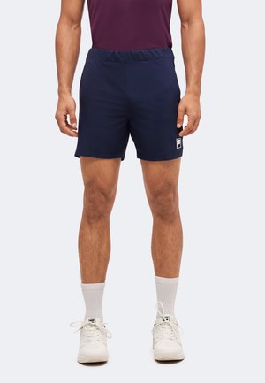 Homme portant un short de sport bleu marine, des chaussettes blanches et des baskets blanches, debout devant un fond uni.
