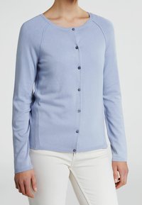 Cardigan bleu clair en tissu doux, avec des manches longues, un col rond, et une fermeture à boutons à l'avant avec des boutons foncés.