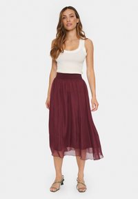 Fustă lungă, burgundy, dintr-un material ușor, cu un braț elastic; asortată cu un top alb, strâns pe corp și sandale cu curele.