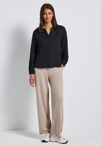 Donna in piedi che indossa una blusa nera a maniche lunghe, pantaloni beige a gamba larga e sneaker bianche su uno sfondo bianco uniforme.