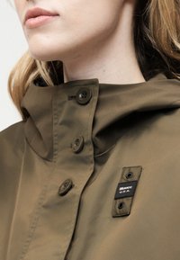 Gros plan sur une personne portant un manteau vert olive à col montant avec une étiquette noire portant l'inscription "Blauer U.S.A." près des boutons.