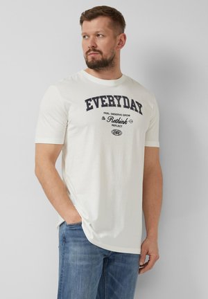 Brodaty mężczyzna w białej koszulce z napisem „EVERYDAY Rethink” i niebieskich dżinsach, stoi z jedną ręką w kieszeni, patrząc w lewo.