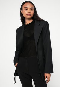 Must mustriga bleiser mustreeritud krae, sile tekstuur, kaks eesmist taskut, kantud läbi sarnaste mustade toppide, koos mustade pükstega.