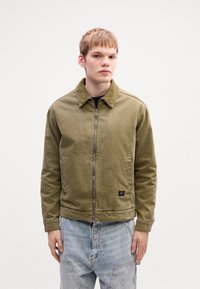Young man wearing olive green zip-up jacket and light blue distressed jeans, standing against plain white background.