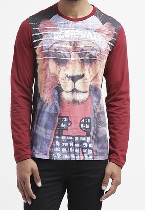 Uomo che indossa una maglia bordeaux a maniche lunghe con una grande immagine di un leone con occhiali da sole, un berretto e abbigliamento a strati sul davanti.