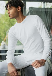 Hvid bluse med lange ærmer lavet af blødt stof, med rund hals og tætsiddende design. Modellen sidder ned og viser outfitets klassiske stil.