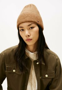 Braune Strickmütze mit gerippter Textur, spitzem Oberteil und kleiner Logoplatte, kombiniert mit einer grünen Jacke und einem hellen Pullover darunter.
