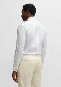 White dress shirt featuring a stand collar, long sleeves with button cuffs, and a smooth texture, paired with light-colored trousers.
