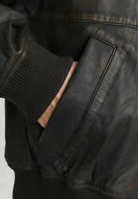 Giubbotto bomber in pelle nera con polsini a costine, pelle testurizzata e una tasca laterale per riporre oggetti. Finitura liscia con una leggera lucentezza.