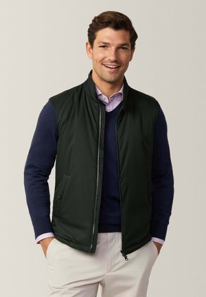 Chaleco acolchado verde oscuro con cuello alto, cierre de cremallera y bolsillos laterales, superpuesto a una sudadera azul marino y una camisa clara con cuello.