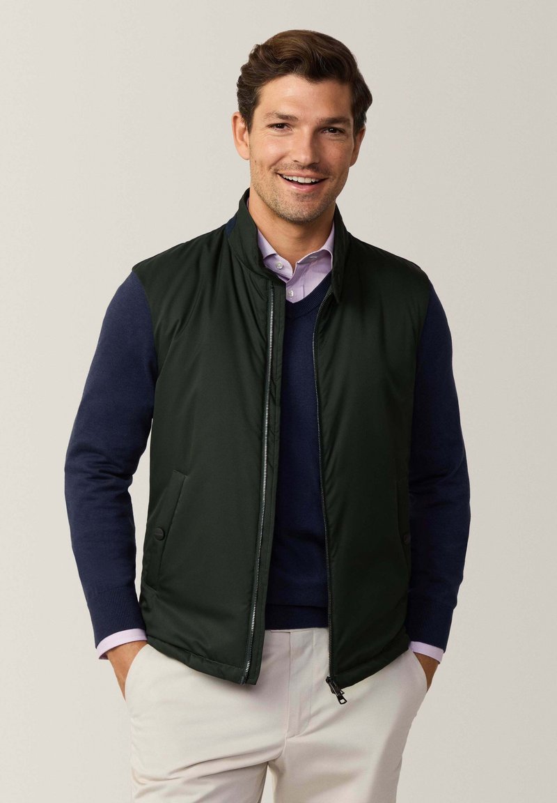 Gilet trapuntato verde scuro con colletto alto, chiusura con zip e tasche laterali, indossato sopra un maglione blu navy e una camicia chiara con colletto.