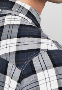 Chemise à carreaux bleue, noire et blanche avec col, présentant un tissu texturé et des détails de couture visibles sur l'épaule et le col.