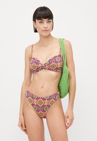 Twee-delige badmode met een paars en oranje patroon, met een geknoopt bovenstuk en lage slipjes, gecombineerd met een groene geweven strandtas.