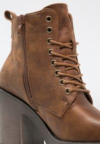 Brązowy sztuczny skórzany botek do kostki z okrągłym noskiem, zamkiem błyskawicznym z boku i wiązanym fasonem. Posiada dekoracyjne oczka i grubą blokow�ą piętę.