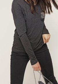 Dunkelgraues Langarmshirt aus weichem Stoff mit einer Tasche aus schwarzen Pailletten, kombiniert mit schwarzen Hosen und einer kleinen Tasche.