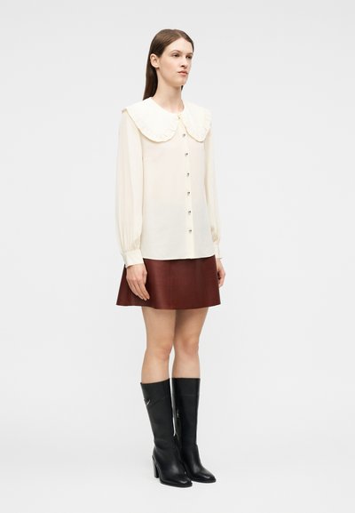 Γυναίκα όρθια, με μια κρεμ μπλούζα με μεγάλο γιακά, καφέ δερμάτινη φούστα και μαύρες μπότες με τακούνι μέχρι το γόνατο, μπροστά σε απλό φόντο.