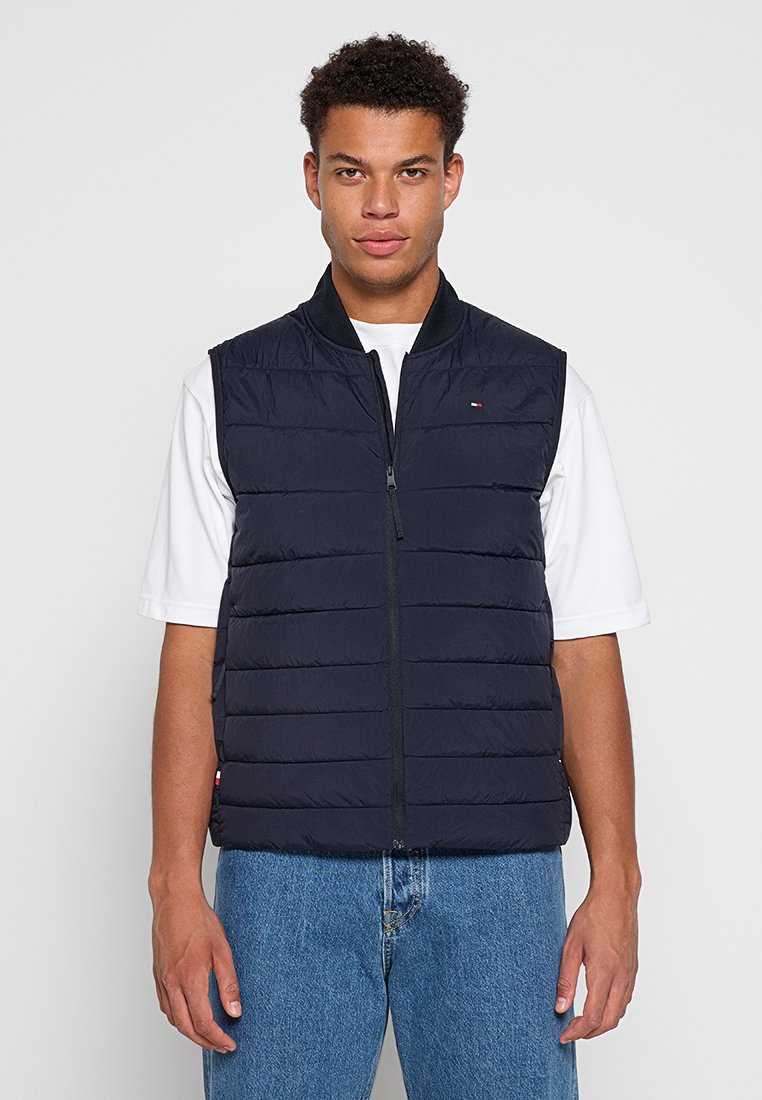 Chaleco sin mangas de plumón azul marino con diseño de acolchado vertical, cierre frontal con cremallera y cuello alto. Llevado sobre una camisa blanca, combinado con jeans azules.