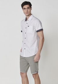 Camisa de botones de manga corta, color gris claro, con un acabado texturizado, cuello negro y detalles en el bolsillo, combinada con pantalones cortos verde oliva arremangados.